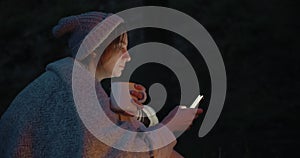 A young woman at dusk sits in a forest opposite a fire and uses a smartphone.