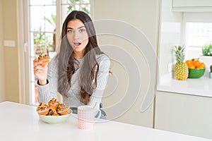 Young woman eating chocolate chips muffins and drinking coffee scared in shock with a surprise face, afraid and excited with fear