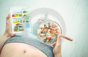 Young woman is eating a fruit salad and using a fitness app on her smartphone after a workout.