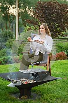 Young woman eating grilled meat sitting at backyard near barbecue