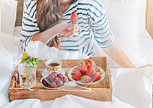 Young woman eating healthy breakfast in bed. Romantic breakfast with strawberries and sweet cherry in a bed