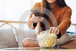 A young woman eating popcorn and searching channel with remote control to watch tv while sitting on sofa at home