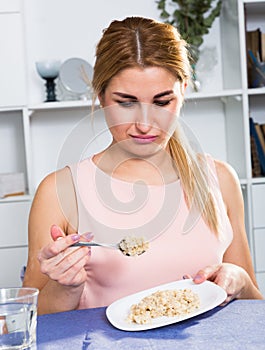 Young woman is eating tasteless oatmeal porridge and feeling disgust