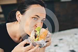 Young woman eating tasty burger in outdoors cafe. Street food festival. Girl love fast food. Unhealthy meal and diet