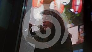 Young woman eats Asian dish sitting at table in dimly lit surroundings of street-side Asian cafe