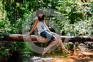 Young woman elegantly sits on a log against the wood and the river.