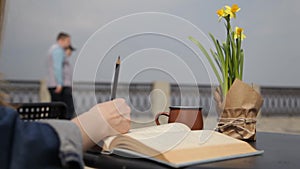 A young woman emphasizes the pencil text in a book sitting at a table in a cafe overlooking the sea.