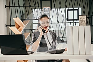 young woman employe sitting at office desk with laptop and talking on phone