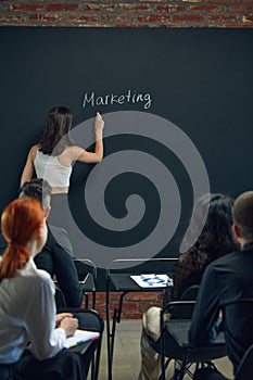 Young woman, employee standing by blackboard and writing word of marketing. Meeting with colleagues. Discussing on