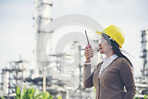 Young woman engineer or construction worker wearing safety hat helmet and holding portable radio with refinery plant background