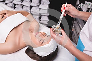 Young woman is enjoying facial procedure at beauty salon. Girl is lying in spa and getting clay mask with pleasure