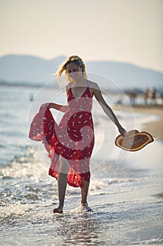 Young woman enjoying her walk on the beach. Beautiful blonde woman in red wavy dress holding straw hat, walking on the shore.