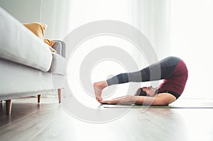 Young Woman enjoying morning yoga exercises doing Halasana pose at home living room near the big window. Active people and healthy