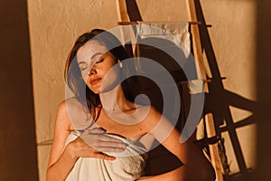 A young woman is enjoying the sun. A beautiful female model wrapped in a white towel stands in the bathroom after a