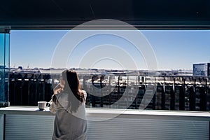 Young woman enjoying view on a sunny day with cup of coffee/tea on balcony in apartment in industrial/business area.Positive young