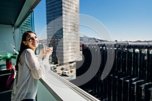 Young woman enjoying view on a sunny day with cup of coffee/tea on balcony in apartment in industrial/business area.Positive young