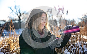 A young woman enjoys a hot drink from a thermos on a walk in winter.