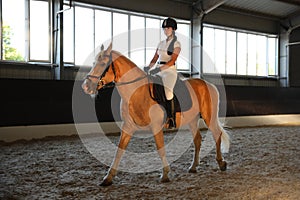 Young woman in equestrian suit riding horse indoors. Beautiful pet