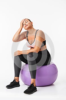 Young woman with excess weight in sporty top and leggings sitting on fitness ball while tiredly closing eyes over white