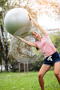 Young woman exercising with pilates ball in the park. Yoga instructor holding fitness ball over her head and training