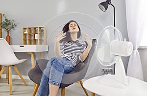 Young woman, exhausted from hot summer weather, sitting by an electric fan at home