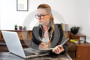 Young woman with eyeglasses, headphones, laptop studying working online writing note pad. Watching online tutorial