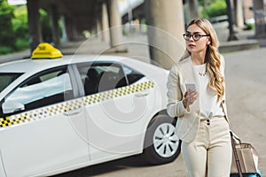 young woman in eyeglasses holding smartphone and looking away while standing near taxi cab