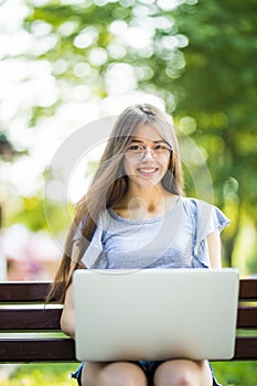 Young woman in eyeglasses sitting on bench in park and using laptop computer