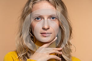 Young woman face with blue eyes, curly natural blonde hair and eyebrows, has no makeup, looking at camera, touching chin. Girl