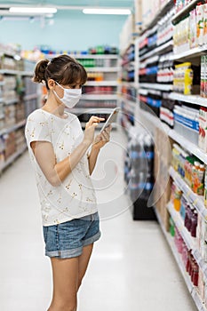 Young woman with face mask using mobile phone and buying groceries in the supermarket