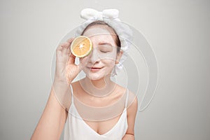 Young woman with facial clay mask holding orange fruit slice covering eyes