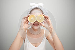 Young woman with facial clay mask holding orange fruit slice covering eyes