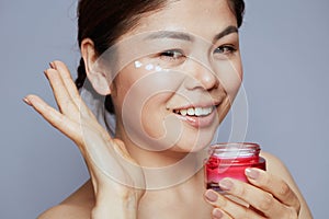 Young woman with facial cream jar and eye cream on face