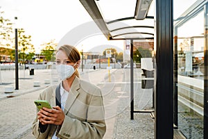 Young woman in facial mask using cellphone standing at bus station