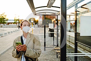 Young woman in facial mask using cellphone standing at bus station