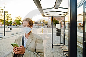 Young woman in facial mask using cellphone standing at bus station