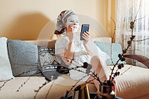 Young woman with facial sheet mask applied looks at smartphone at home after bath relaxing on couch.