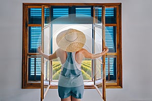 Young woman farmer, dressed in blue jumpsuit shorts, looks out window with open shutters on grape field