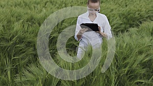 Young woman farmer wearing white bathrobe is checking harvest progress on a tablet at the green wheat field. New crop of