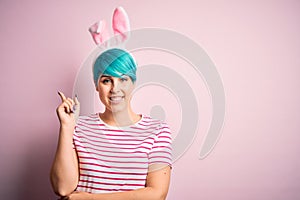 Young woman with fashion blue hair wearing easter rabbit ears over pink background with a big smile on face, pointing with hand