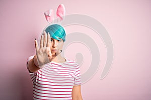 Young woman with fashion blue hair wearing easter rabbit ears over pink background doing stop sing with palm of the hand