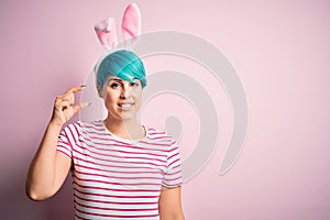 Young woman with fashion blue hair wearing easter rabbit ears over pink background smiling and confident gesturing with hand doing