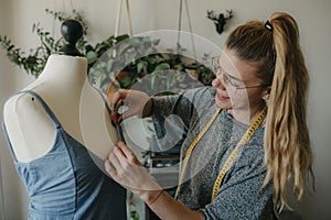 Young woman fashion designer measuring mannequin, seamstress holding tape working with dummy in cozy home or tailor shop,