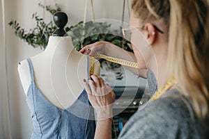 Young woman fashion designer measuring mannequin, seamstress holding tape working with dummy in cozy home or tailor shop,