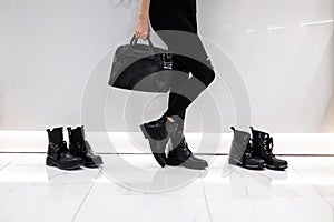Young woman with a fashionable black leather bag in stylish boots in jeans stands near a white wall in a store next to shoes.