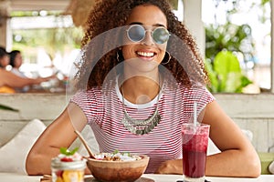 Young woman in fashionable round shades has fresh drink and eats tasty exotic dish, smiles broadly, rests at cafeteria