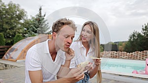Young woman feeding her laughing friend a slice of cheesy pizza near poolside. Couple eating pizza together