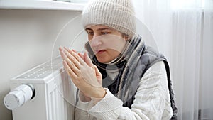 Young woman feeling cold in house sitting at heating radiator. Concept of energy crisis, high bills, broken heating system,