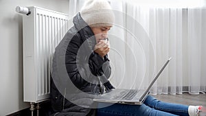 Young woman feeling cold wearing winter hat and coat sitting by the heater nd working on laptop