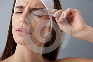 Young woman feeling pain while plucking eyebrows with tweezers